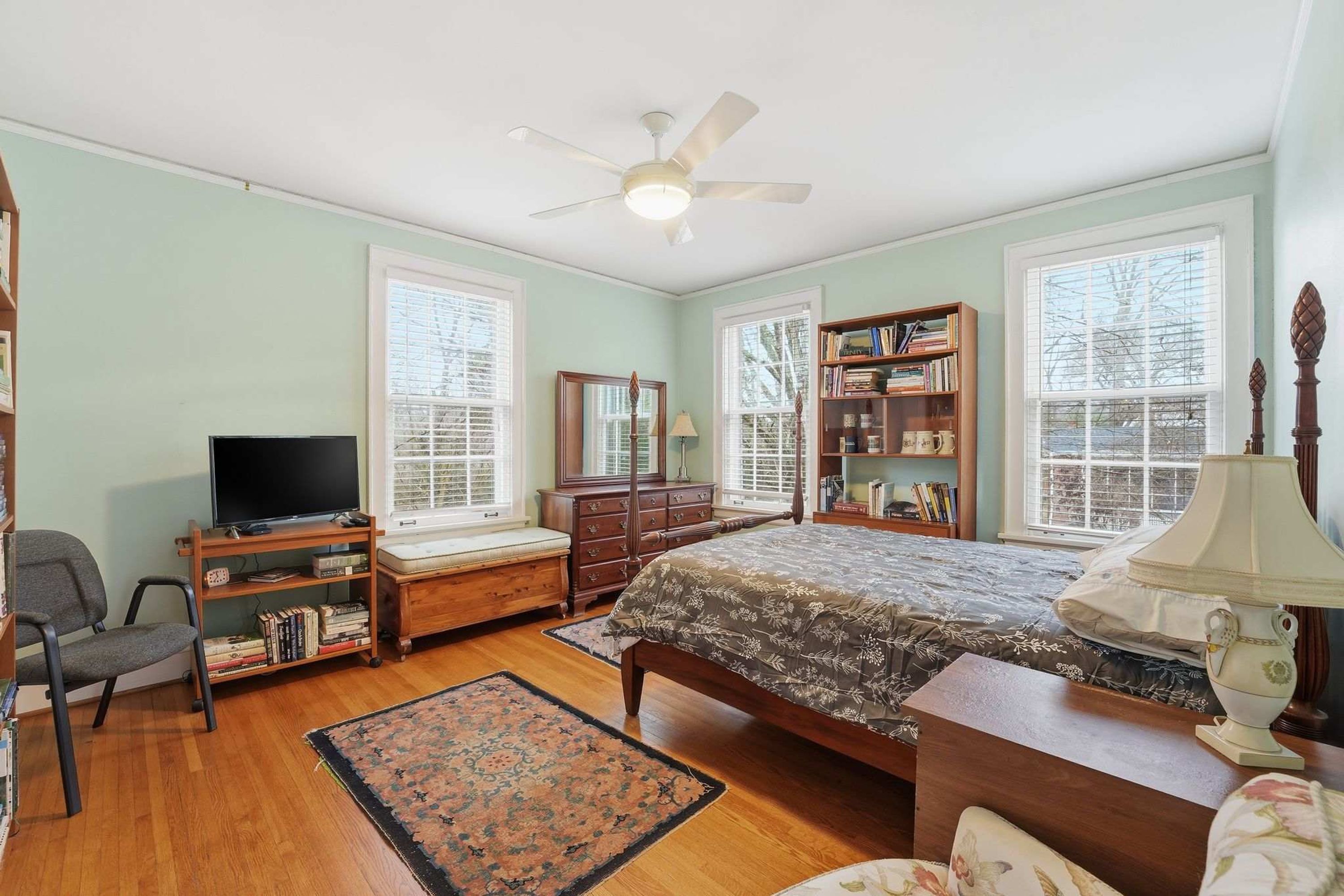 Quarto com cama, escrivaninha, cadeira, televisão, estante de livros e janela com persianas.