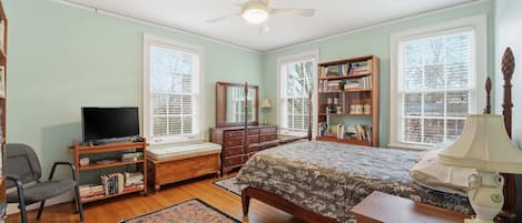 Quarto com cama, escrivaninha, cadeira, televisão, estante de livros e janela com persianas.