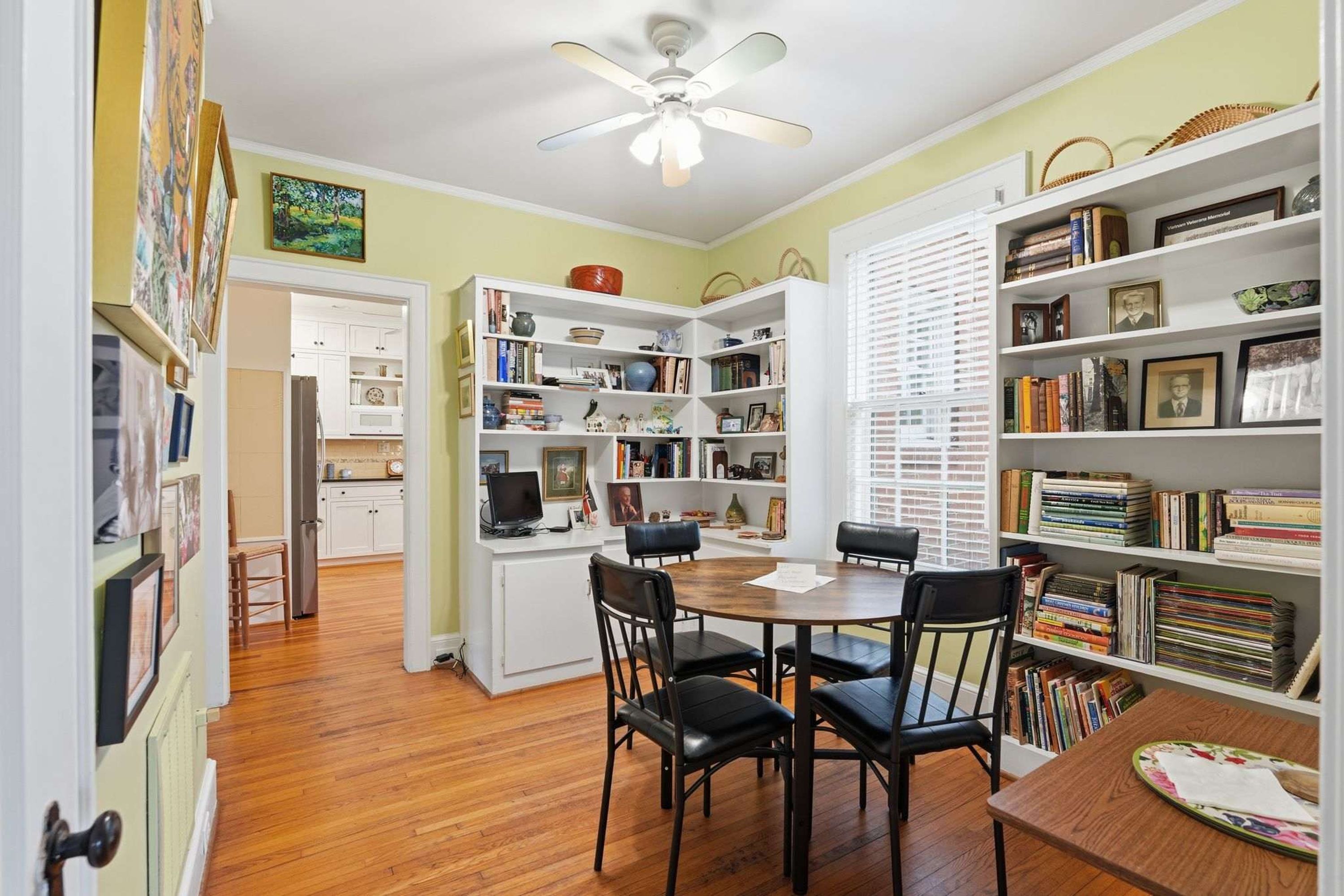A room with a dining table, chairs, a ceiling fan, and bookshelves filled with books and decorative items.