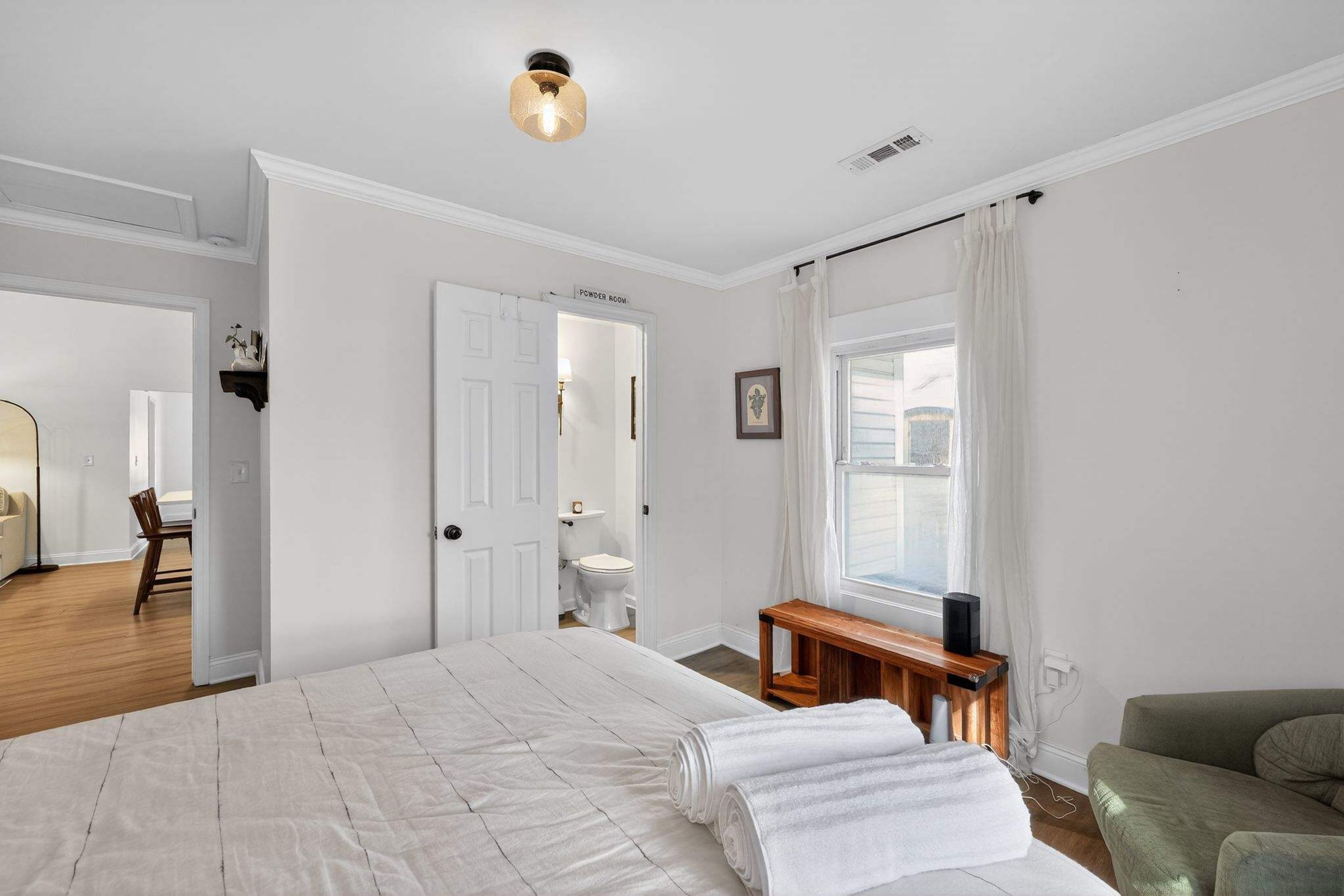 Un dormitorio con una cama grande, una mesita de noche, una silla, una ventana con cortinas y una puerta que da a otra habitación.