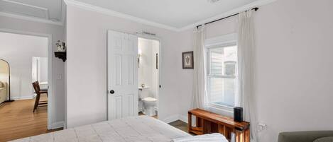 Un dormitorio con una cama grande, una mesita de noche, una silla, una ventana con cortinas y una puerta que da a otra habitación.