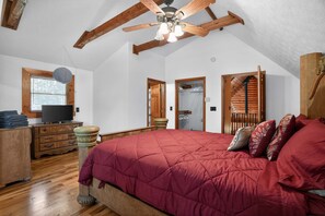 Un dormitorio con una cama grande, vigas de madera, un ventilador de techo, un televisor y una ventana con persianas.