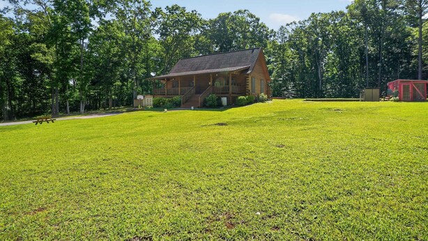 A wooden cabin with a porch, surrounded by a well-maintained lawn and dense trees. - Cabin Outdoor Space Views Gym Game Room (Ellenwood)