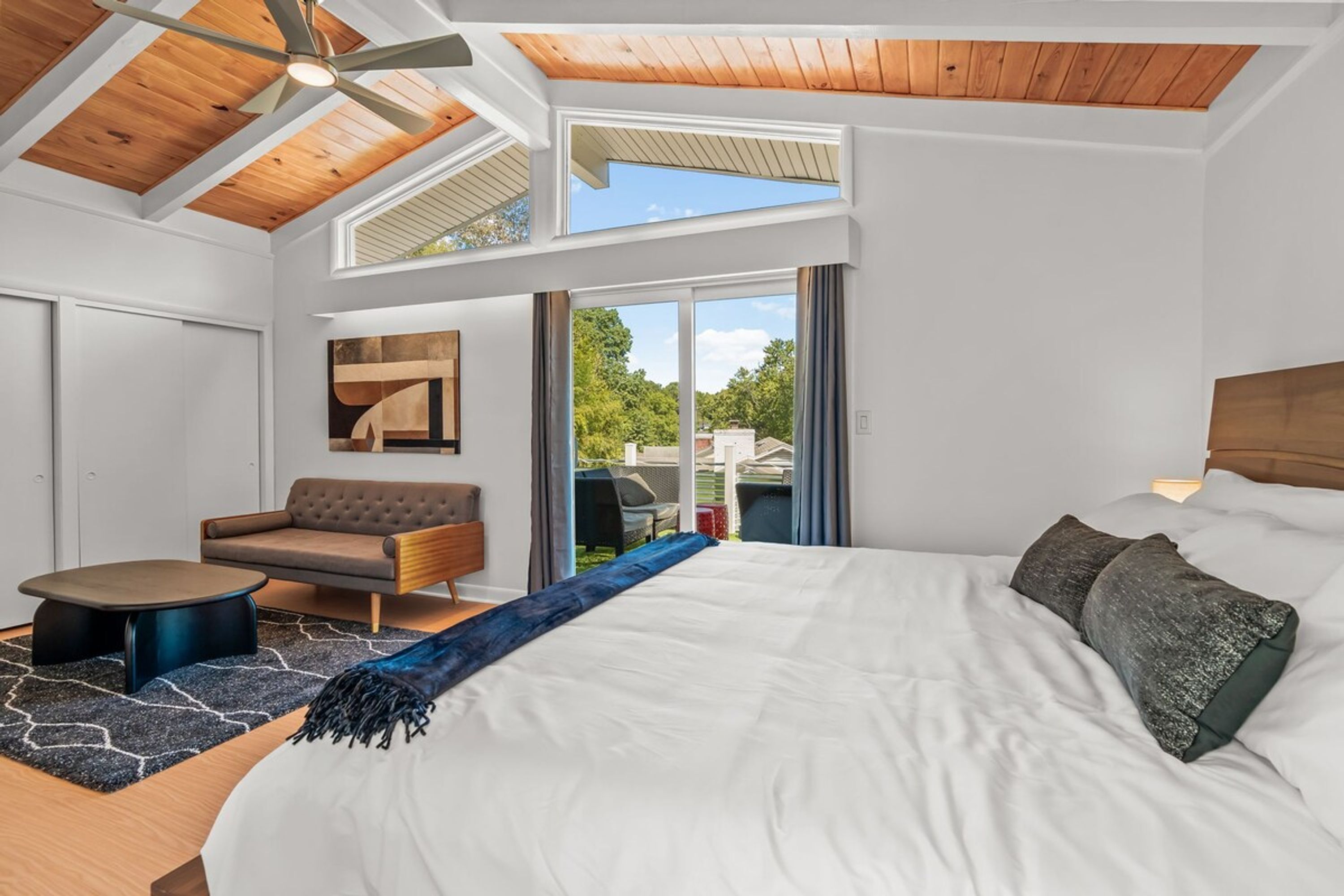 A modern bedroom with a large bed, a sofa, a small table, and a ceiling fan.