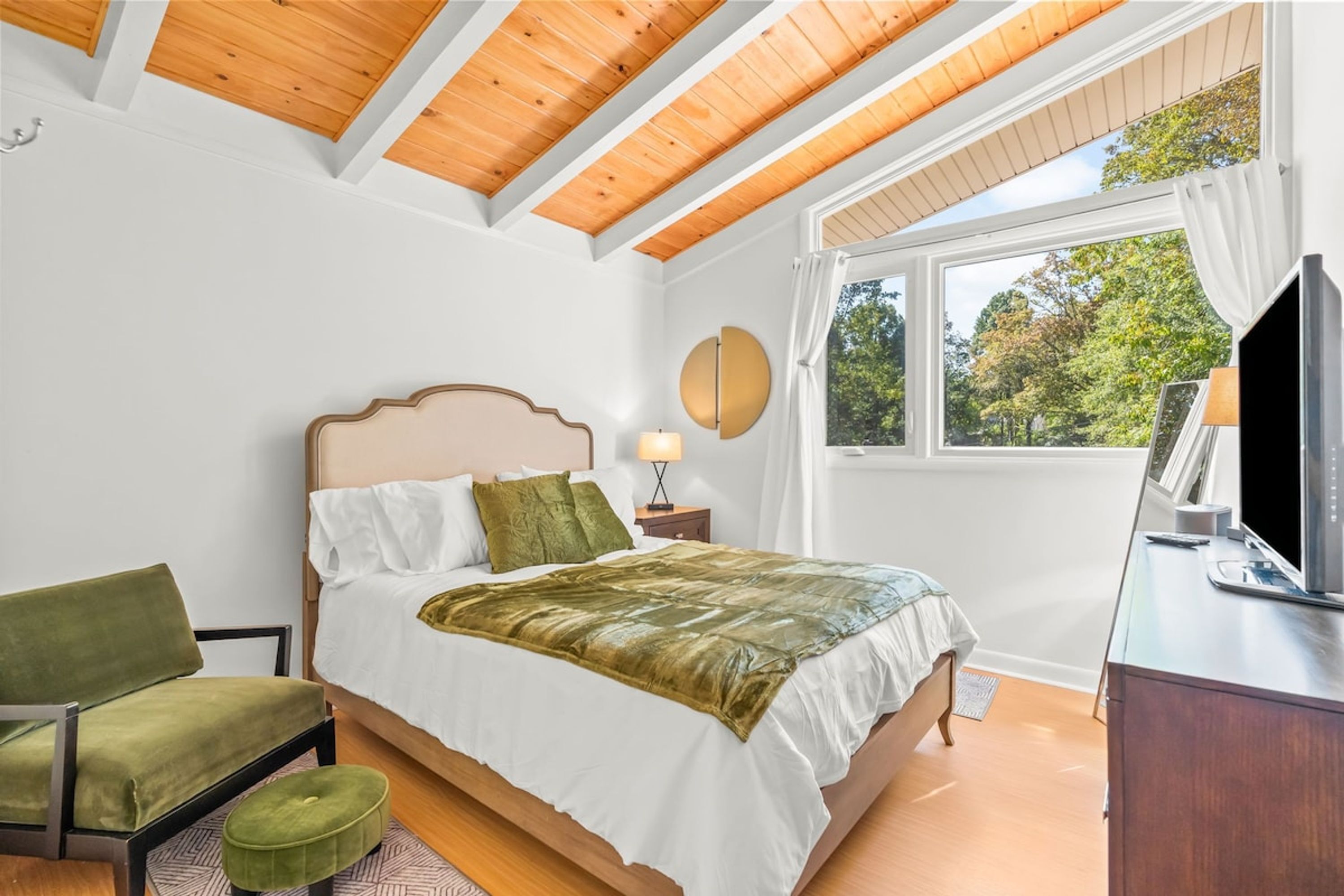 Un dormitorio con cama, una silla, un televisor y una ventana con vistas a árboles.