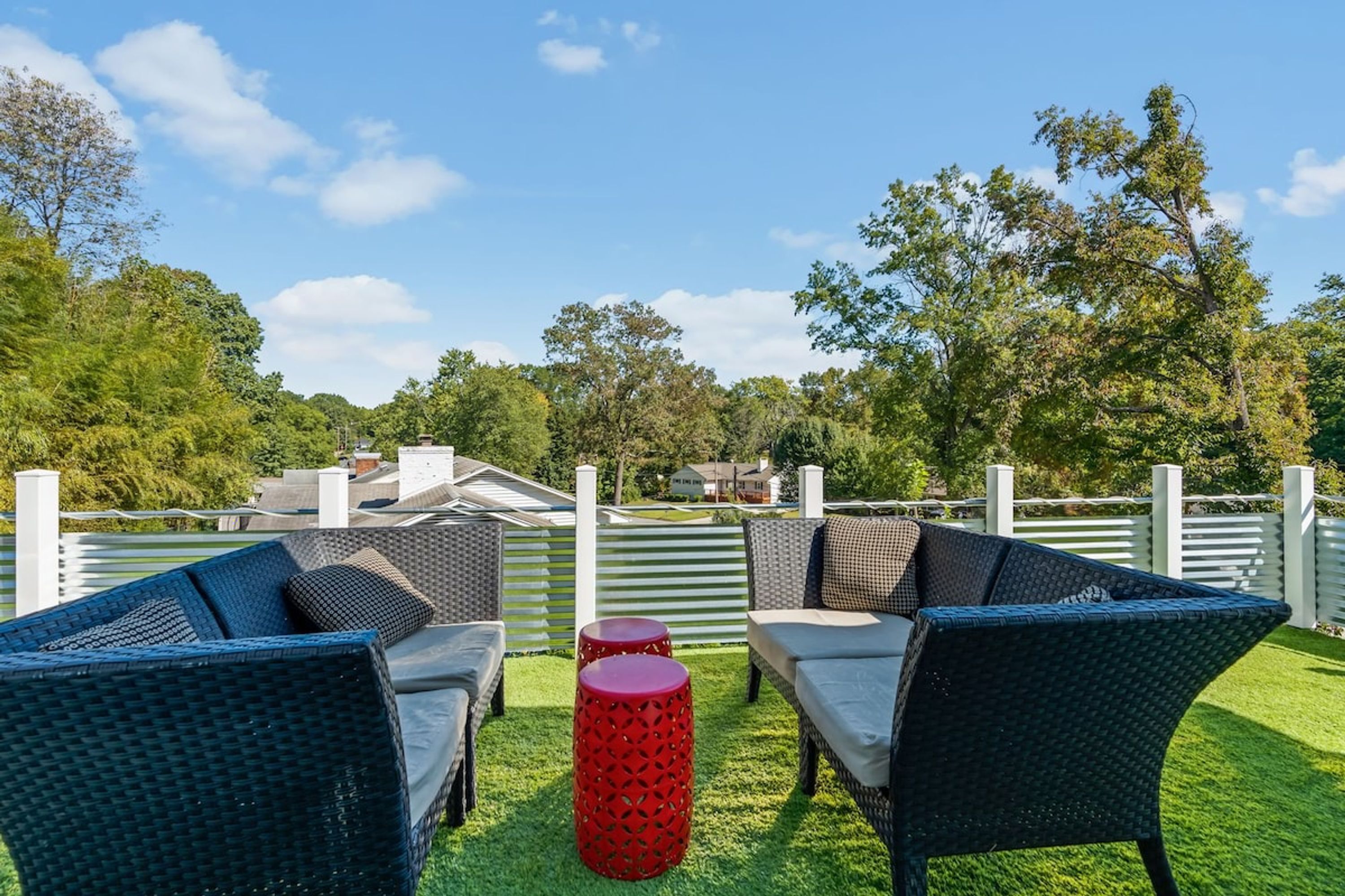 Un espace extérieur aménagé avec des meubles en osier et un tabouret rouge, entouré d’une clôture blanche et de verdure.
