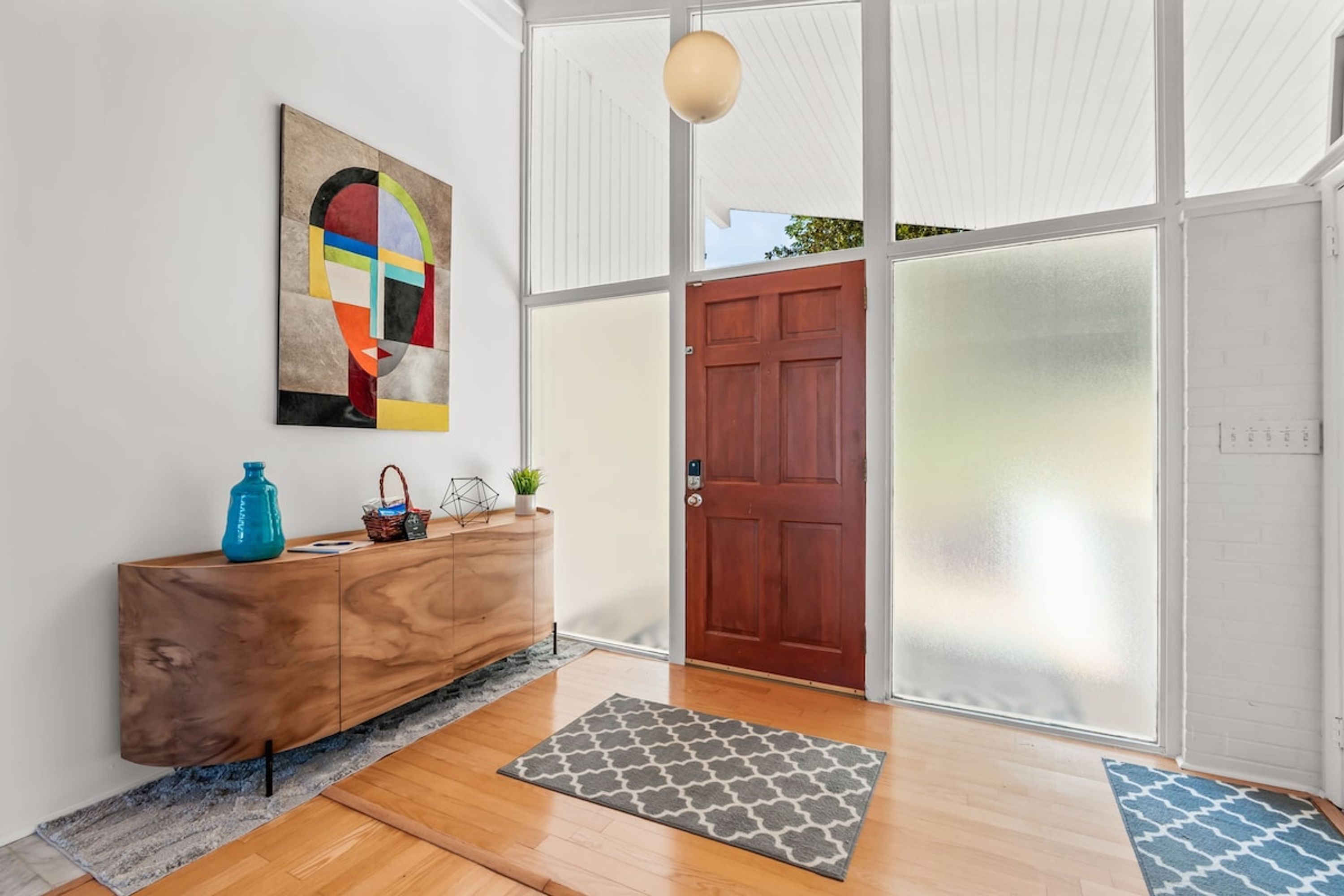 A modern interior with a wooden floor, a grey patterned rug, a wooden console, a colorful abstract painting, and a wooden door.