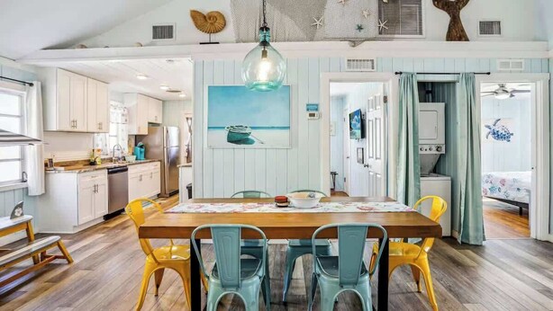 A kitchen with a wooden table and chairs, a hanging light fixture, and a painting on the wall. - Beach Life 3 - Sunshine Lane (Treasure Island)