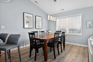 A dining room with a wooden table and chairs, framed pictures on the wall, and a large window with blinds. - Coastal 4BR Retreat Hot tub Water Views Deck Pet-friendly (Holly Ridge)