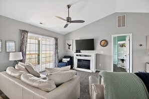 A living room with a sofa, a flat-screen TV, a fireplace, and a ceiling fan. - Coastal 4BR Retreat Hot tub Water Views Deck Pet-friendly (Holly Ridge)