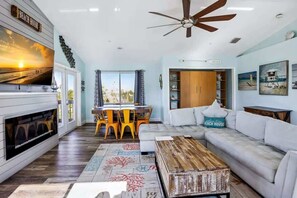 A modern living room with a fireplace, a ceiling fan, and a dining area with yellow chairs. - The Gulf House | Walk to Beach | 5BR Retreat (Treasure Island)