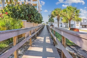 A wooden boardwalk leading to a building with balconies, surrounded by greenery and palm trees. - Beach Life 2 - Sunshine Lane (Treasure Island)