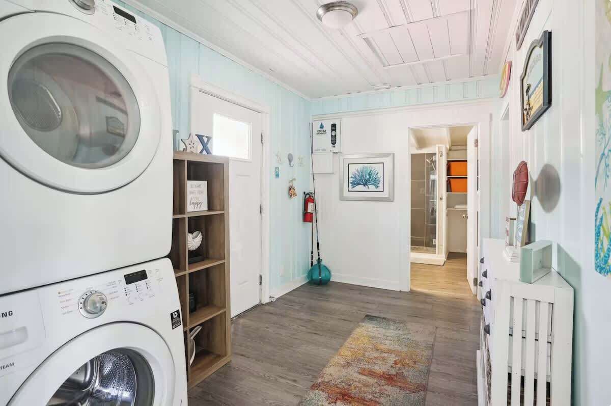 Ett tvättrum med en tvättmaskin, en torktumlare och en hylla med hopvikta kläder. Det finns en dörr som leder till ett annat rum, en brandsläckare och en tavla som hänger på väggen.