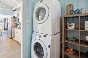 A laundry area with stacked washing machines, a wooden shelf with decorative items, and a kitchen area in the background. - Beach Life 2 - Sunshine Lane (Treasure Island)