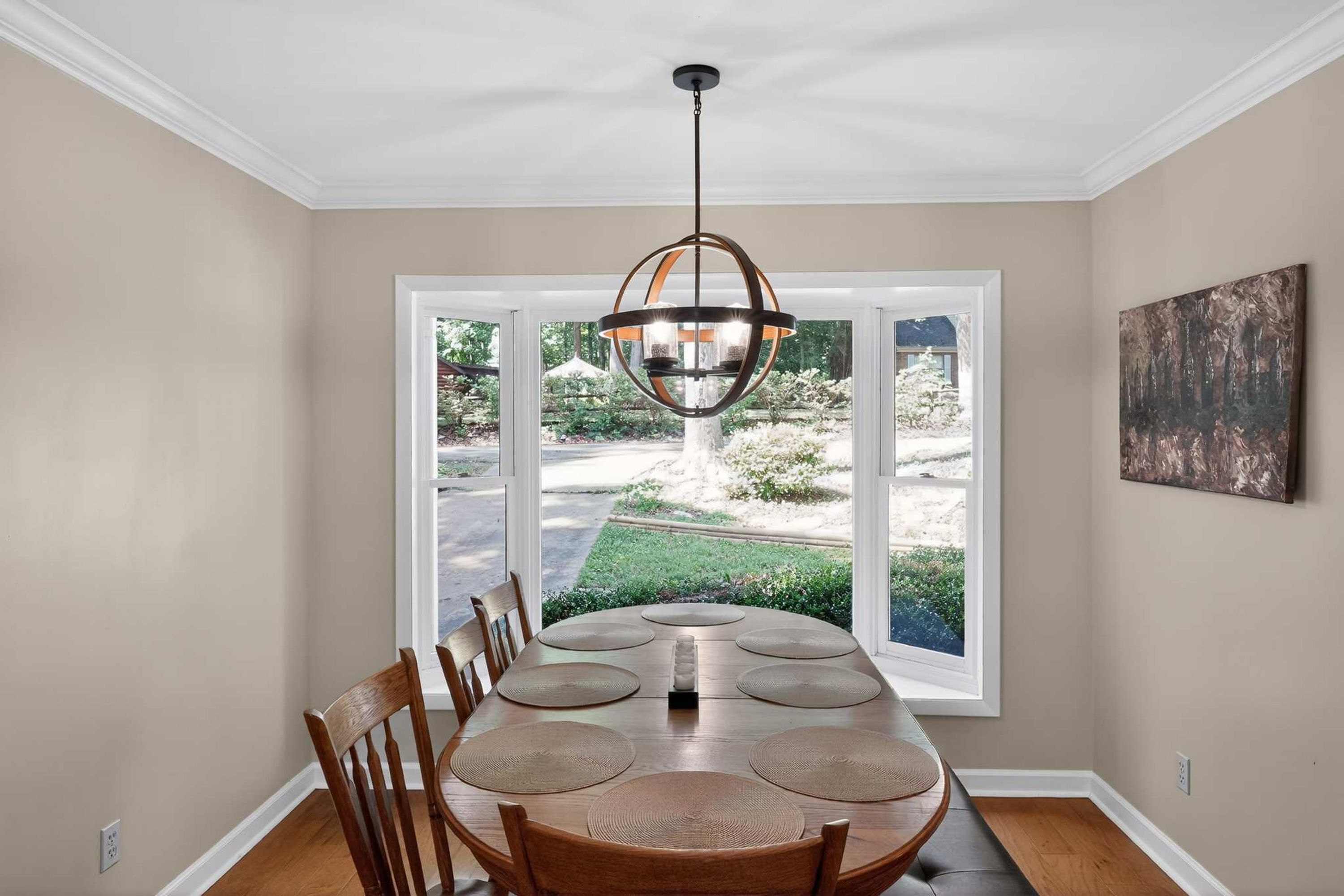 A dining area with a large table, chairs, a hanging light fixture, and a view through large windows.