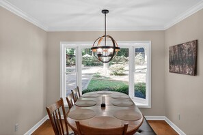 A dining area with a large table, chairs, a hanging light fixture, and a view through large windows.