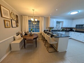 A modern kitchen with white cabinetry, a dining area with a round table and chairs, and a large window with curtains.