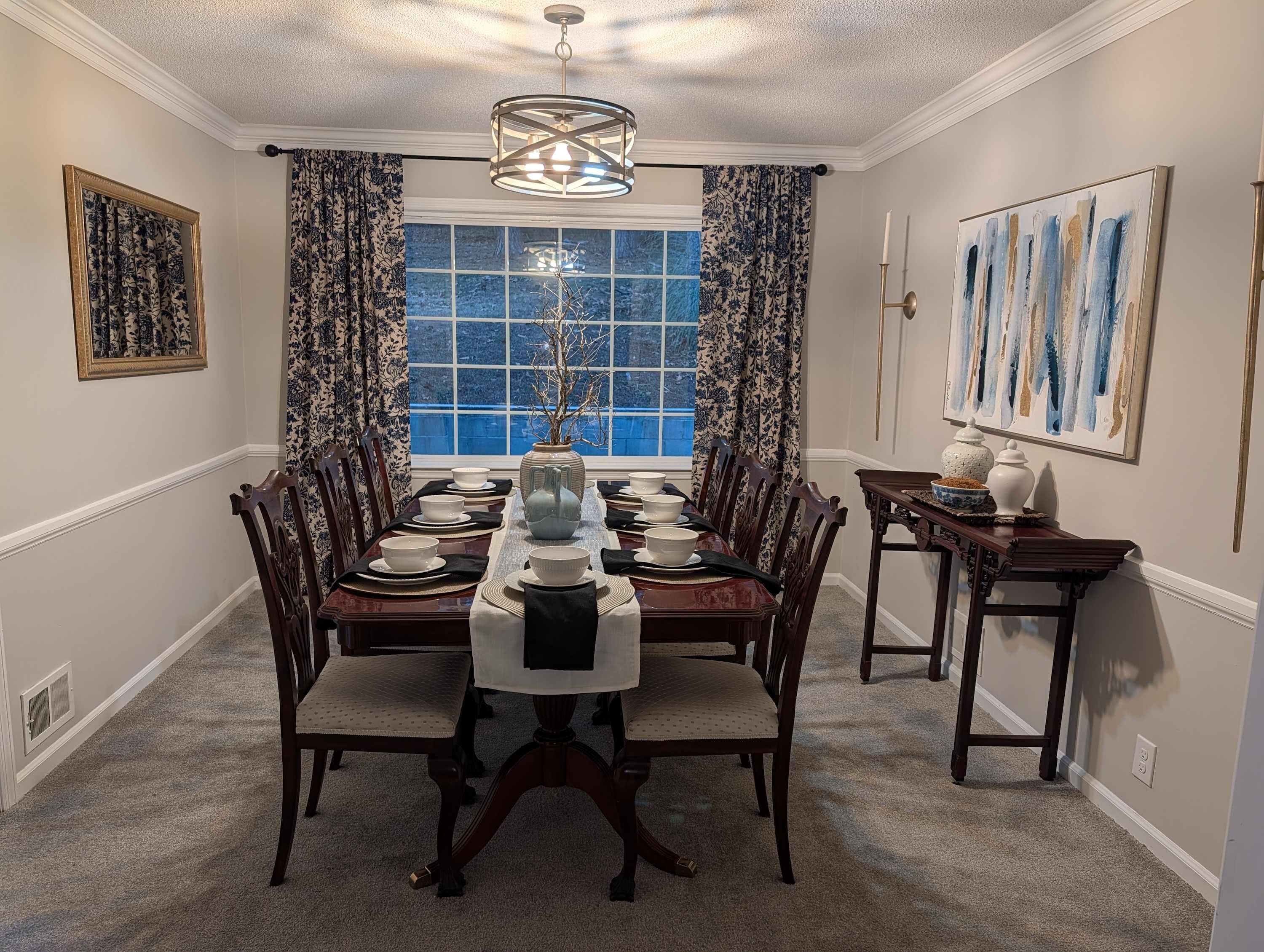 A dining room with a long table set for six, flanked by chairs, a vase centerpiece, and a window with curtains.