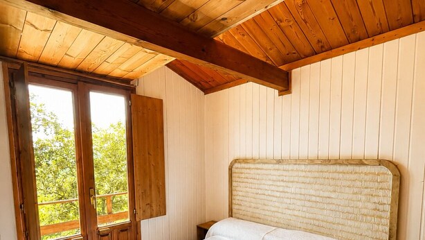 Un dormitorio con techo de madera, cama con colcha blanca con textura y una ventana con contraventanas de madera.