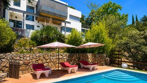 Piscina con sillas de descanso y sombrillas, rodeada por un muro de piedra y follaje.