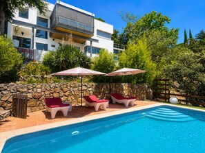 A swimming pool with lounge chairs and umbrellas, surrounded by a stone wall and greenery.