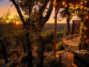 A wooden pergola with string lights, a hanging chair, and a view of a natural landscape during sunset.