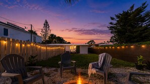 Un patio trasero con un hogar para fogata, mobiliario exterior, un cerco de madera y vistas a una masa de agua al atardecer.
