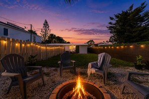 Ein Garten mit Feuerstelle, Sitzgelegenheiten im Freien, Holzzaun und Blick auf ein Gewässer in der Abenddämmerung.