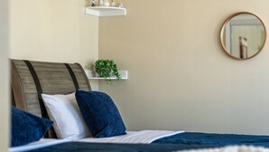 Un dormitorio con una cama, almohadas, una planta en la mesita de noche, un espejo y una repisa con objetos decorativos.