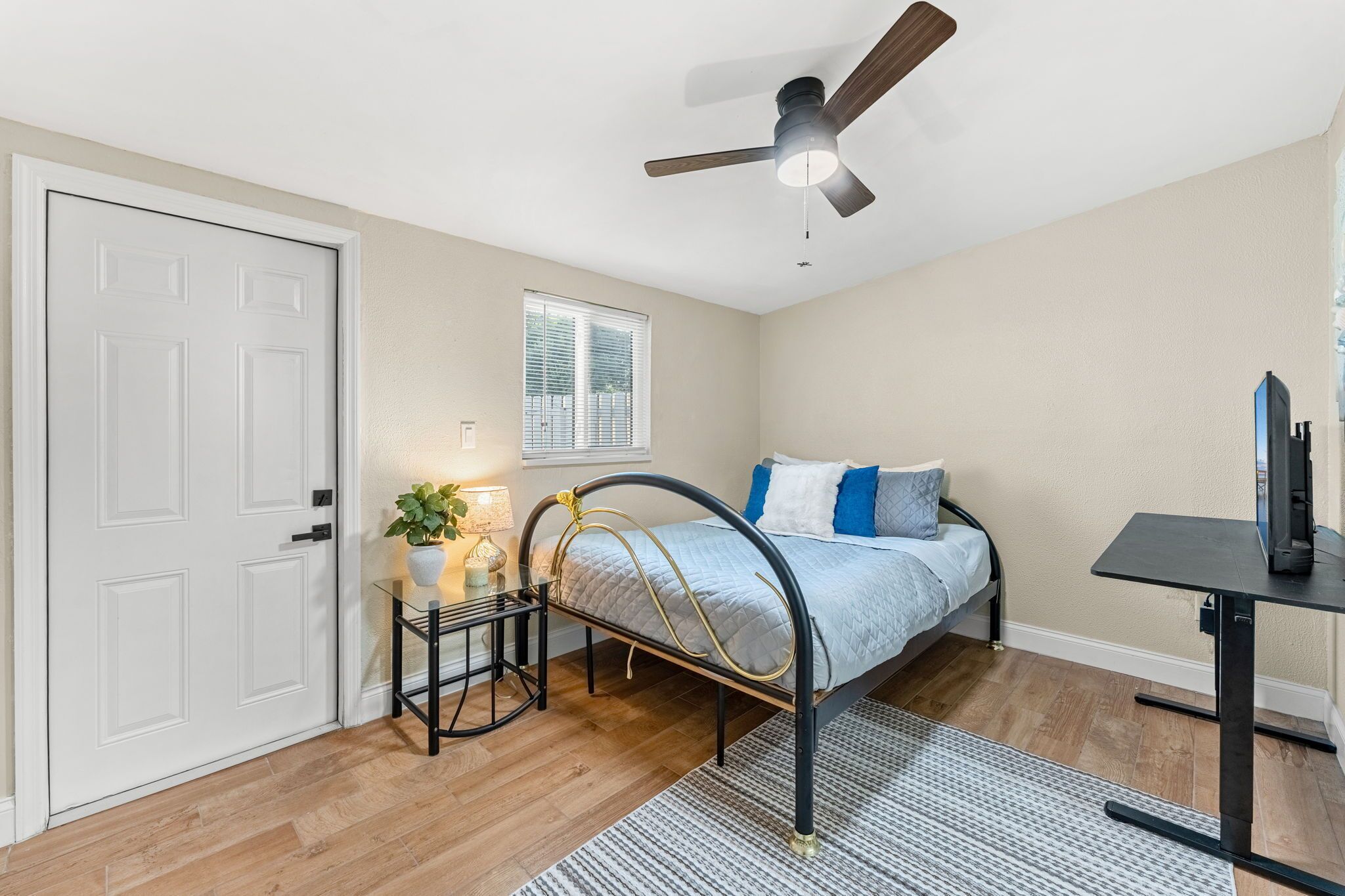 A bedroom with a bed, bedside table, lamp, ceiling fan, window, and a desk with a computer.