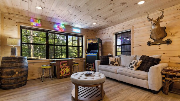 Ein gemütliches Blockhaus-Interieur mit Holzbalkendecke, einer Couch mit Waldkissen, einer Neonreklame und einem aufgehängten Hirschkopf.