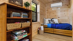 Ein gemütliches Zimmer mit einer Holzbücherreih, einem Bett mit blauer Decke und einem Fenster mit Klimaanlage.