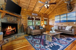 A cabin interior with a stone fireplace, leather sofas, a wooden coffee table, and a large painting of a mountain landscape. - Mountain View Cabin | Theatre & Game Room | Hot Tub (Blue Ridge)