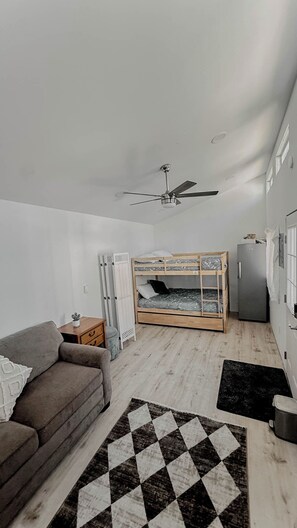 A room with a bunk bed, a sofa, a ceiling fan, a refrigerator, and a small table with a plant.