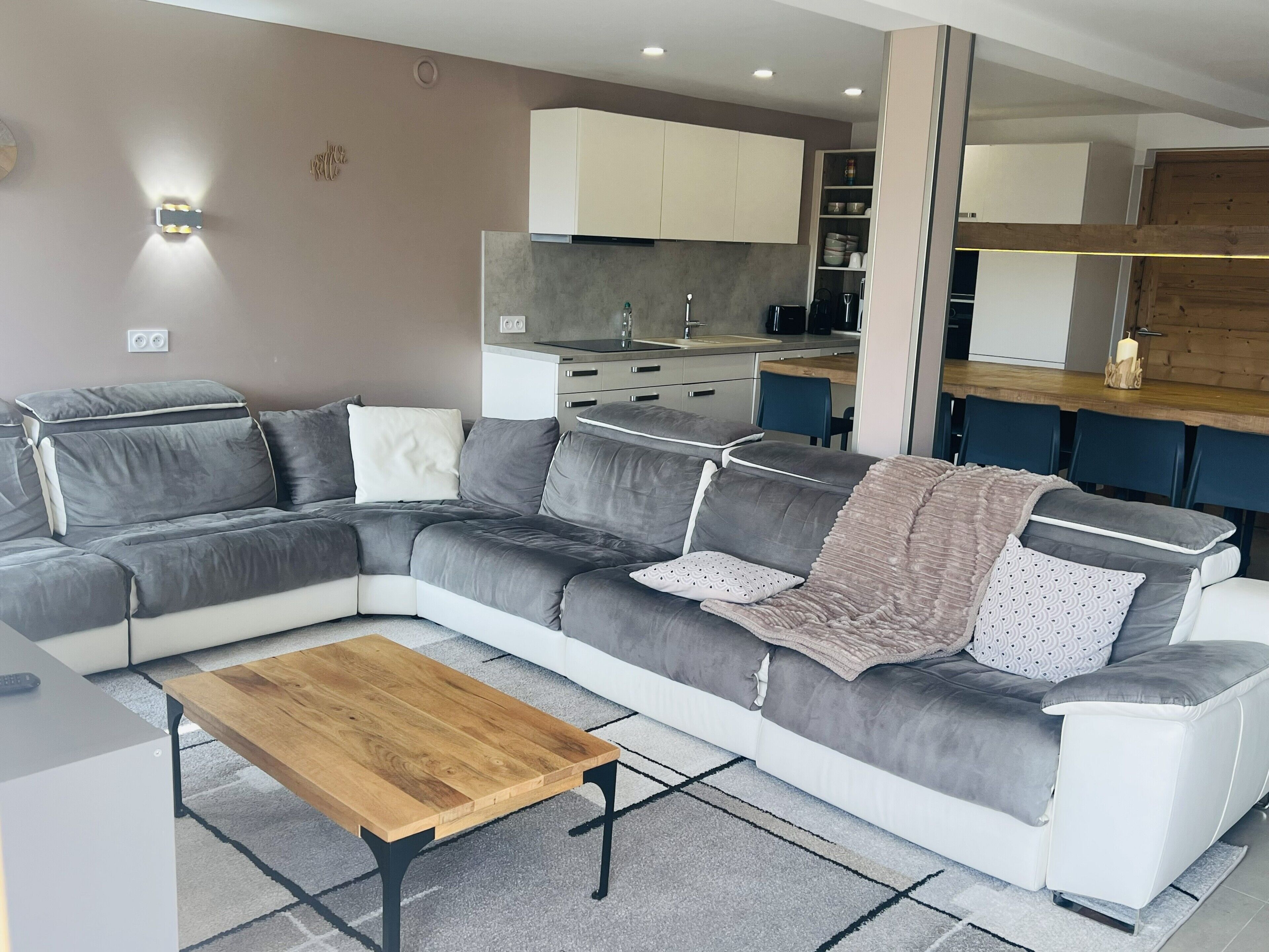 Una sala de estar moderna con un sofá modular, una mesa de centro de madera y una cocina al fondo.