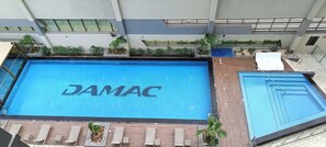 Ein großer, überdachter Pool mit dem Schriftzug 'DAMAC' auf der Wasseroberfläche, umgeben von Liegestühlen und Topfpflanzen.