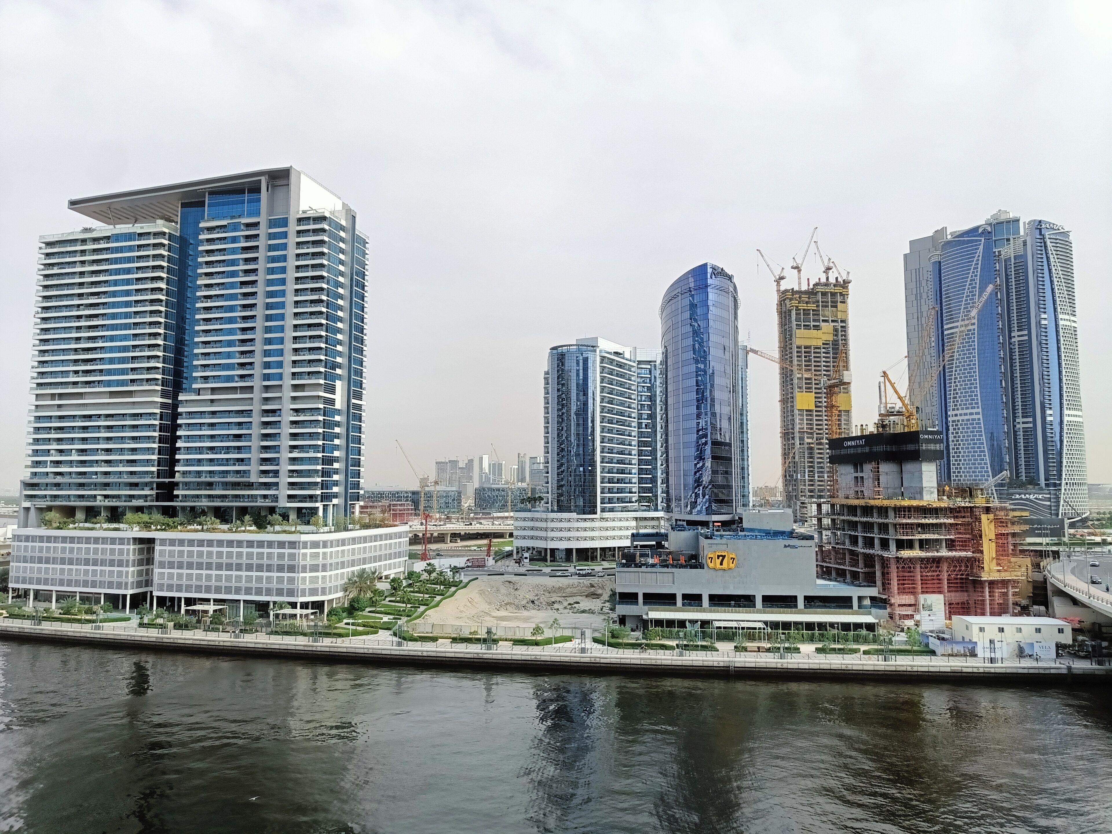A modern cityscape with high-rise buildings, a waterfront, and construction cranes.