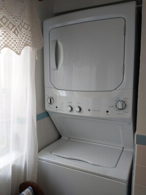 Una lavadora de carga frontal con perillas de control y puerta transparente, ubicada en un baño con pared de azulejos blancos y una ventana con cortina translúcida.