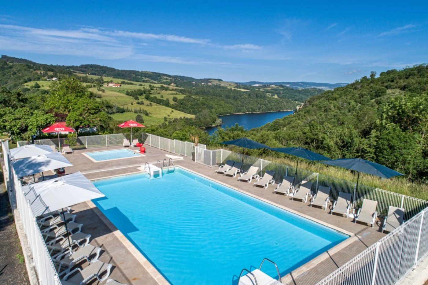 Une piscine rectangulaire, bordée de chaises longues et d’ombrelles, entourée d’une clôture blanche, et offrant une vue sur un lac et des collines en arrière-plan.