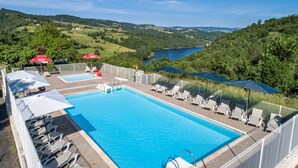 Uma piscina retangular com espreguiçadeiras e guarda-sóis, cercada por uma vedação branca, com vista para um lago e colinas ao fundo.