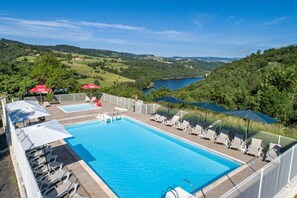 Une piscine rectangulaire, bordée de chaises longues et d’ombrelles, entourée d’une clôture blanche, et offrant une vue sur un lac et des collines en arrière-plan.
