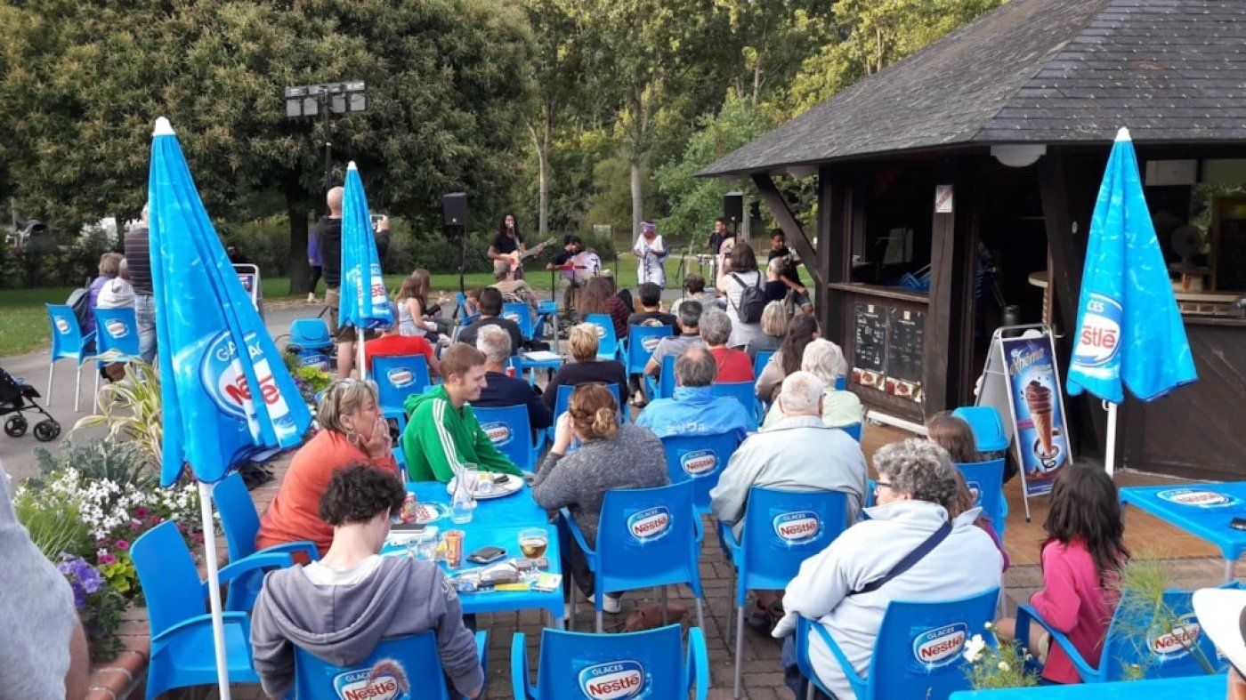 Een terras met blauwe stoelen en parasols, een podium met muzikanten, en een klein gebouw op de achtergrond.
