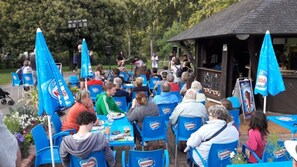 An outdoor seating area with blue chairs and umbrellas, a stage with musicians, and a small building in the background.