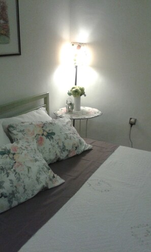 Een bed met bloemenbeddengoed, een nachtkastje met een lamp, en een wandcontactdoos.