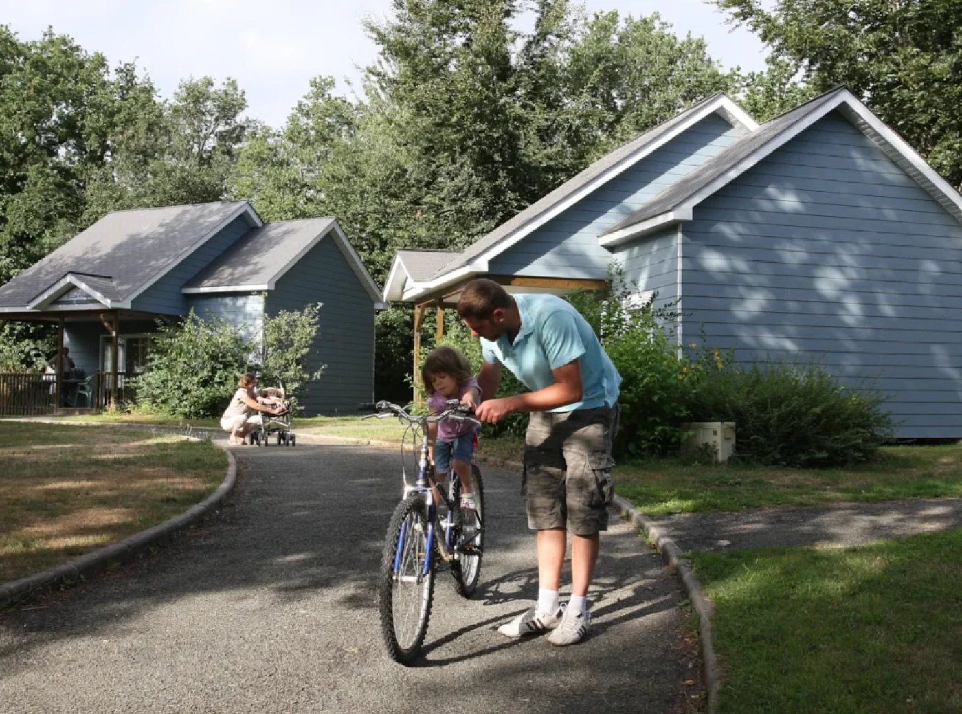 Een woonwijk met huizen, een verharde oprit en een persoon die een kind met een fiets helpt.