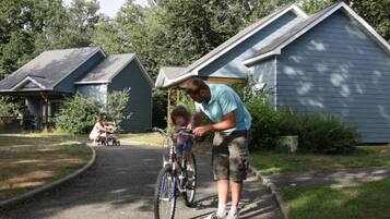 Een woonwijk met huizen, een verharde oprit en een persoon die een kind met een fiets helpt.