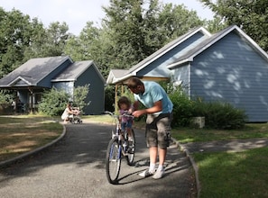 A residential area with houses, a paved driveway, and a person assisting a child with a bicycle.