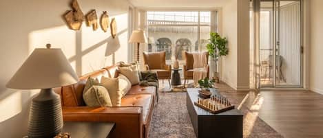 A modern living room with a sofa, coffee table, and a chessboard. There is a window with a view of a bridge, and a sliding glass door leading to an outdoor area.