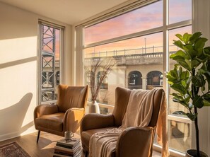 A room with two leather armchairs, a large window with a view of a bridge, a potted plant, and a stack of books. - Luxury 2BR Apartment Balcony, Gym & Parking (Cleveland)
