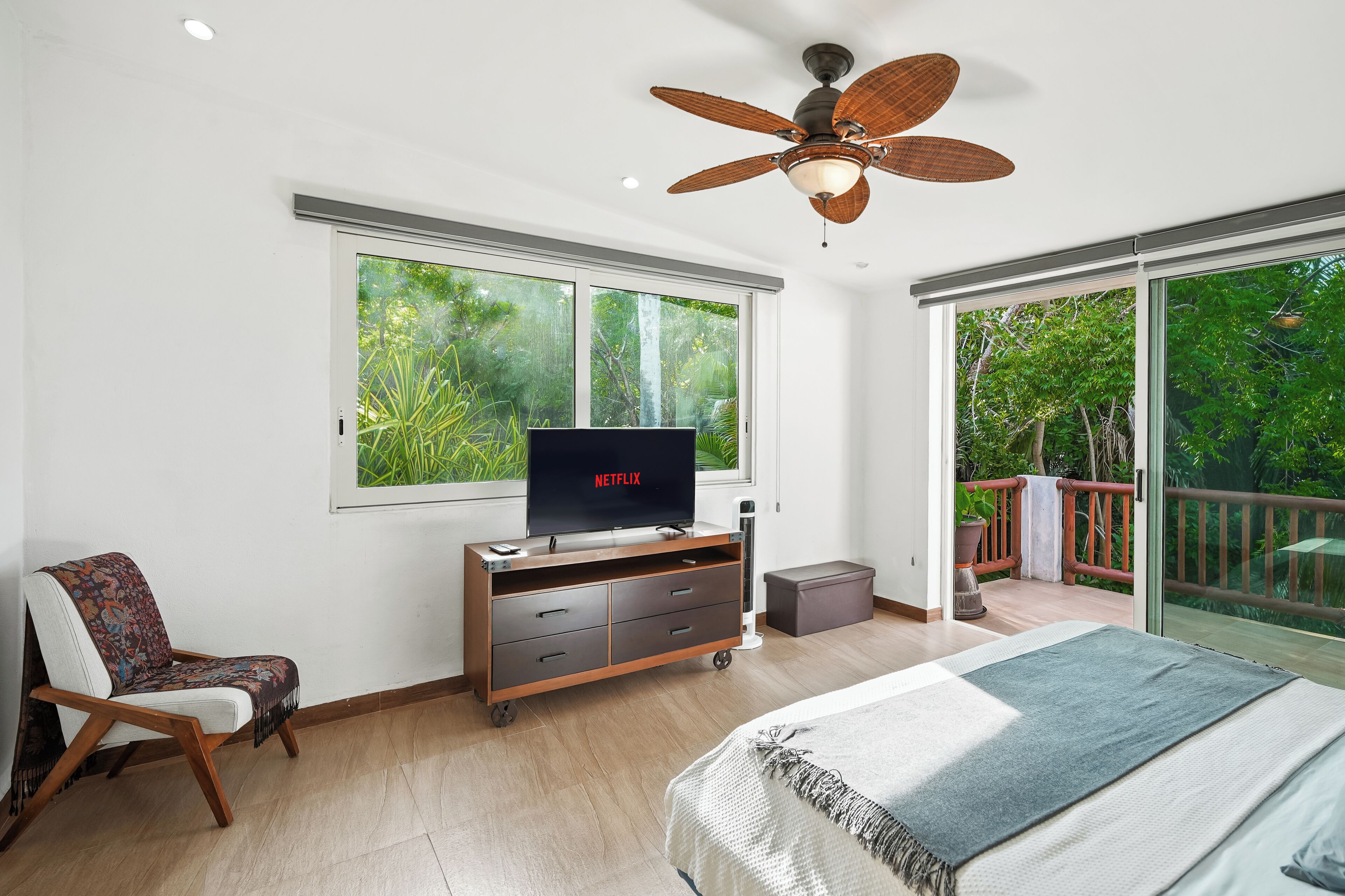 A bedroom with a bed, a TV, a ceiling fan, a chair, a dresser, and a sliding glass door leading to a balcony.